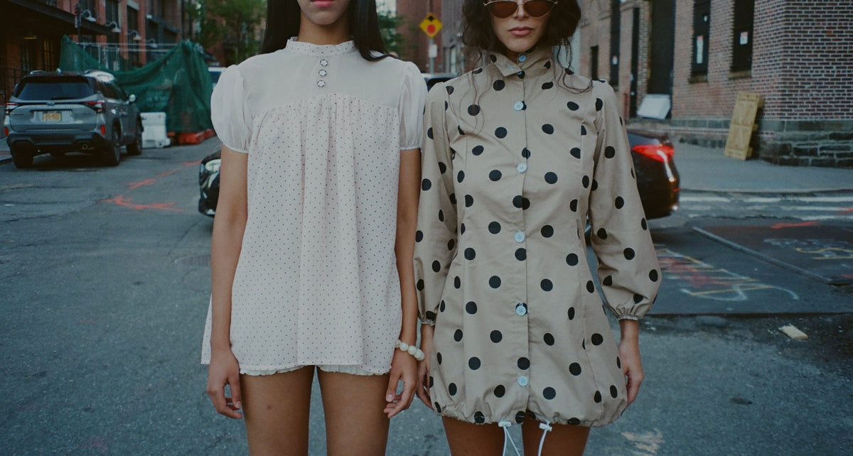 Two women standing on a city street wearing short polka-dot dresses. The woman on the left wears a white dress with small black dots, and the woman on the right wears a beige dress with large black polka dots.